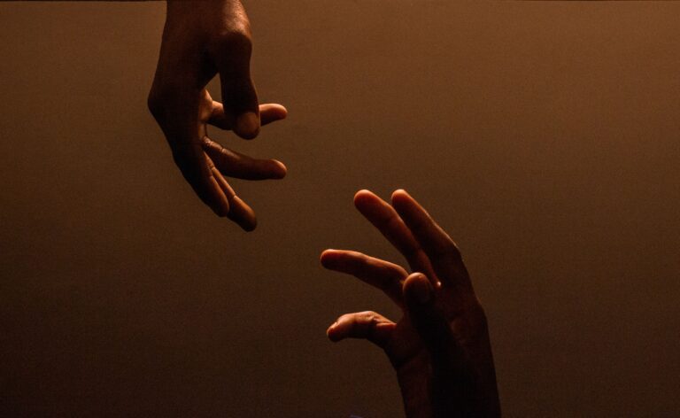 Two hands reaching towards each other in warm, low light against a dark brown background, with space between their fingertips suggesting connection, support and hope.