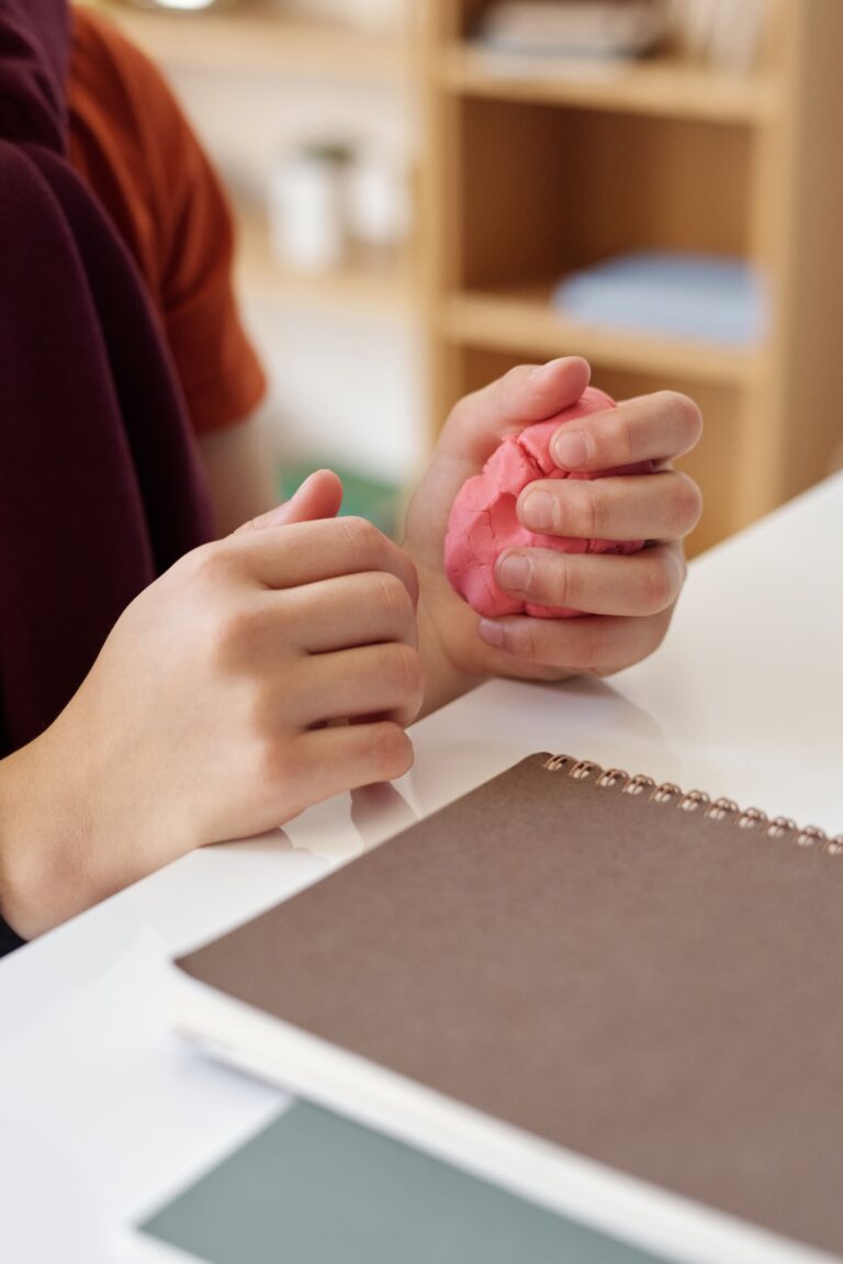 A person in a burgundy top gently kneads bright pink modelling clay at a white table, with a notebook and tablet nearby in a softly lit home setting.