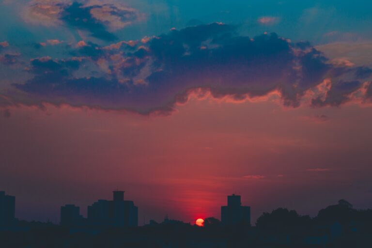 A vivid red sun sets behind a dark city skyline, beneath dramatic blue-grey clouds glowing with pink and coral light.