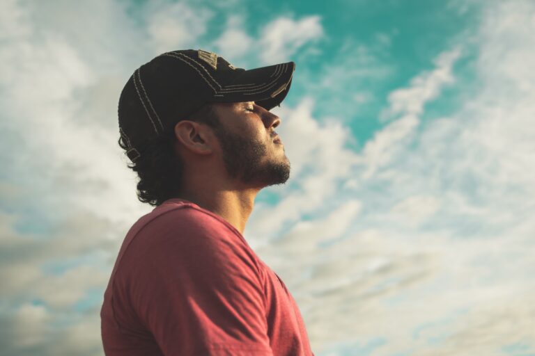 Man in a black cap and red shirt looking upward beneath a blue sky with soft clouds.